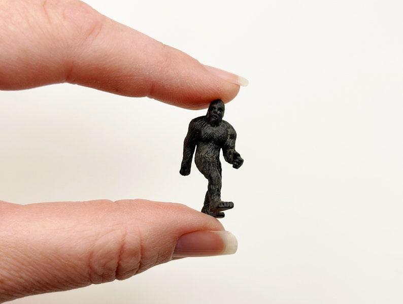 May include: A small black plastic figurine of a Bigfoot or Sasquatch walking, held between two fingers.