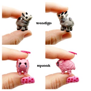 May include: Four miniature figurines are displayed. Two are gray and white creatures labeled "wendigo," and two are pink creatures labeled "squonk." The figurines are held between fingers against a white background. The "squonk" figurines have blue tear drops.