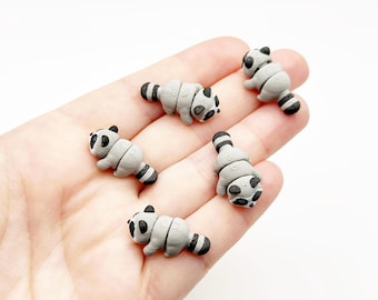 Op de afbeelding: Vijf kleine, grijs en zwarte wasbeerfiguurtjes met getextureerde oppervlakken. Elke miniatuurwasbeer heeft een afgerond lichaam, een gestreepte staart en een opvallend zwart maskerpatroon rond de ogen, afgewerkt met een matte finish.