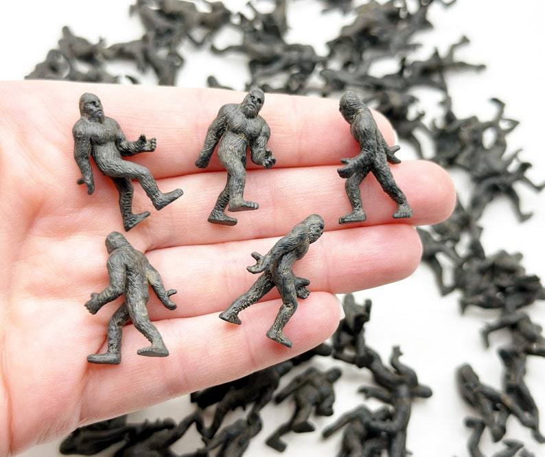 May include: A hand holding five small, gray plastic figurines of Bigfoot. The figurines are all in different poses and are about 2 inches tall.