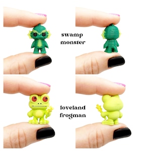 May include: Miniature green Swamp Monster and Loveland Frogman figurines. The Swamp Monster has a textured head and fins. The Loveland Frogman is lime green with red eyes and a yellow star. Text reads "swamp monster" and "loveland frogman".