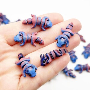 May include: A collection of small, colorful, hand-painted, ceramic,  lizard-shaped beads. The beads are a mix of blue and pink with a swirl pattern.