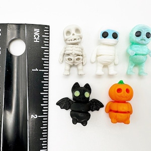 Peut inclure: Cinq petites figurines sur le th&egrave;me d'Halloween, dont un squelette blanc, une momie blanche, un personnage bleu clair translucide, une chauve-souris noire aux yeux brillants et une citrouille orange, &agrave; c&ocirc;t&eacute; d'une r&egrave;gle noire de 5 centim&egrave;tres.