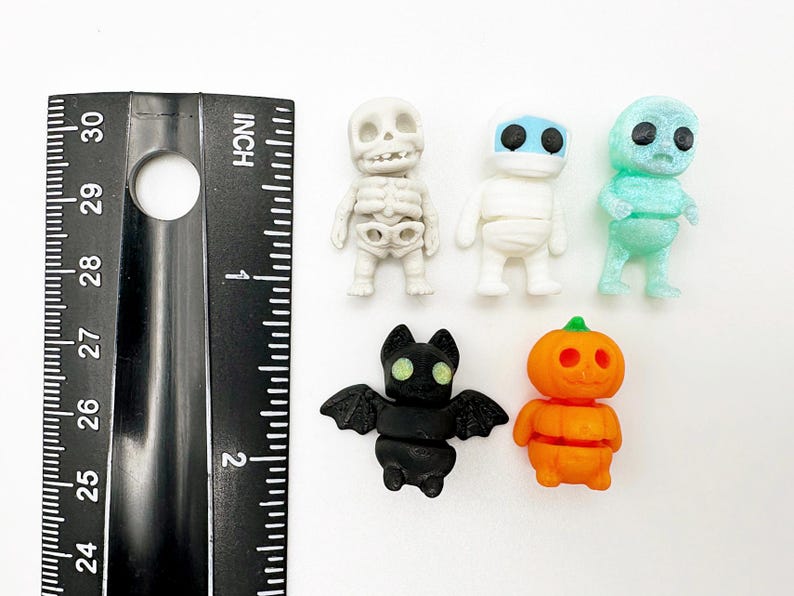 Peut inclure: Cinq petites figurines sur le th&egrave;me d'Halloween, dont un squelette blanc, une momie blanche, un personnage bleu clair translucide, une chauve-souris noire aux yeux brillants et une citrouille orange, &agrave; c&ocirc;t&eacute; d'une r&egrave;gle noire de 5 centim&egrave;tres.