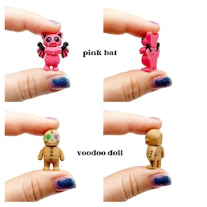 Peut inclure: Une figurine miniature de chauve-souris rose avec des ailes noires et une figurine de poup&eacute;e vaudou beige avec des d&eacute;tails cousus. La chauve-souris rose a de grands yeux et des oreilles pointues, tandis que la poup&eacute;e vaudou pr&eacute;sente une bouche cousue.