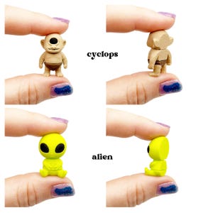 Peut inclure: Une figurine miniature de cyclope beige avec un &oelig;il noir et un pagne marron, et une figurine miniature d'extraterrestre jaune fluo avec de grands yeux noirs. Le cyclope est vu de face et de dos, l'extraterrestre de face et de profil.