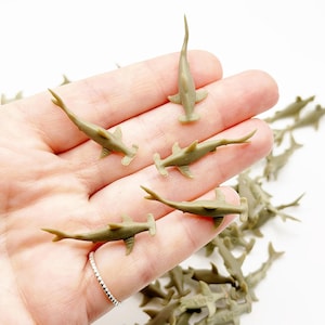 May include: A collection of small, olive-green hammerhead shark figurines. Several are arranged in a person's open palm, with others scattered in the background. The sharks have a distinctive hammer-shaped head and a long, slender body.