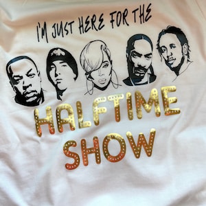 Op de afbeelding: Wit T-shirt met een zwarte afbeelding van vijf muzikanten en de tekst "I'm just here for the halftime show" in gouden glitterletters.