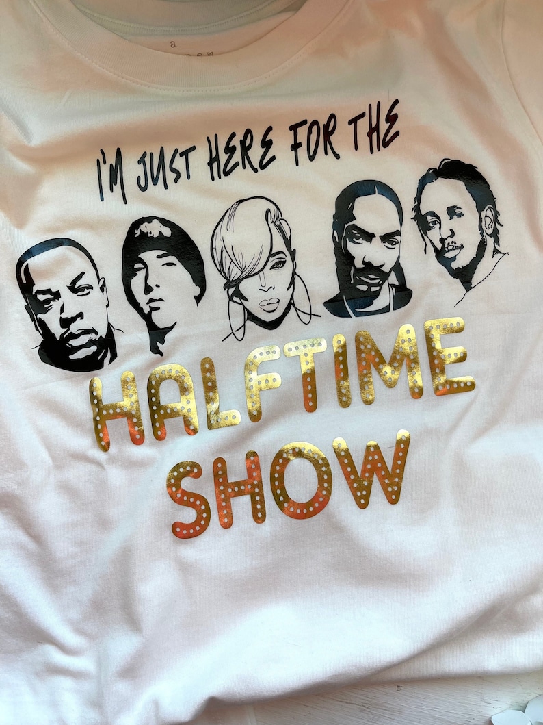 Op de afbeelding: Wit T-shirt met een zwarte afbeelding van vijf muzikanten en de tekst "I'm just here for the halftime show" in gouden glitterletters.
