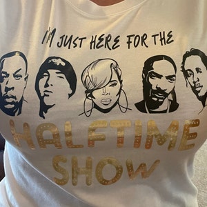 Op de afbeelding: Wit T-shirt met zwarte tekst die "I'm just here for the halftime show" luidt. De tekst wordt omringd door zwart-witte afbeeldingen van vier mensen.