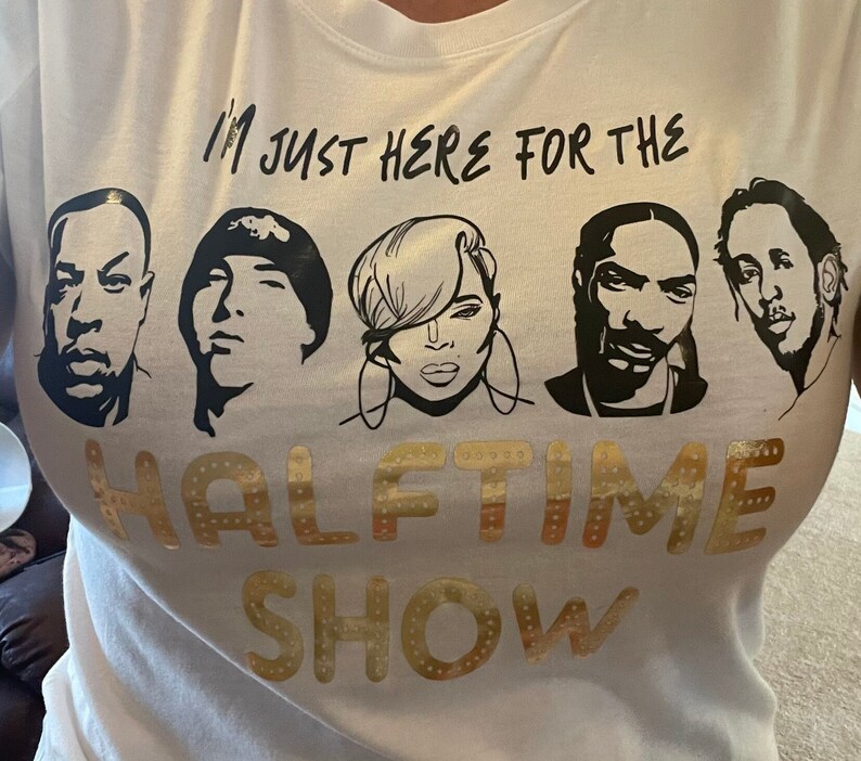 Op de afbeelding: Wit T-shirt met zwarte tekst die "I'm just here for the halftime show" luidt. De tekst wordt omringd door zwart-witte afbeeldingen van vier mensen.