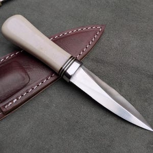 Pu&ograve; includere: Un coltello artigianale con un manico colorato chiaro e una lama argentata, appoggiato su un fodero in pelle marrone. Il manico ha dettagli argentati. Il fodero &egrave; cucito con filo bianco. Il coltello &egrave; progettato per l'uso.