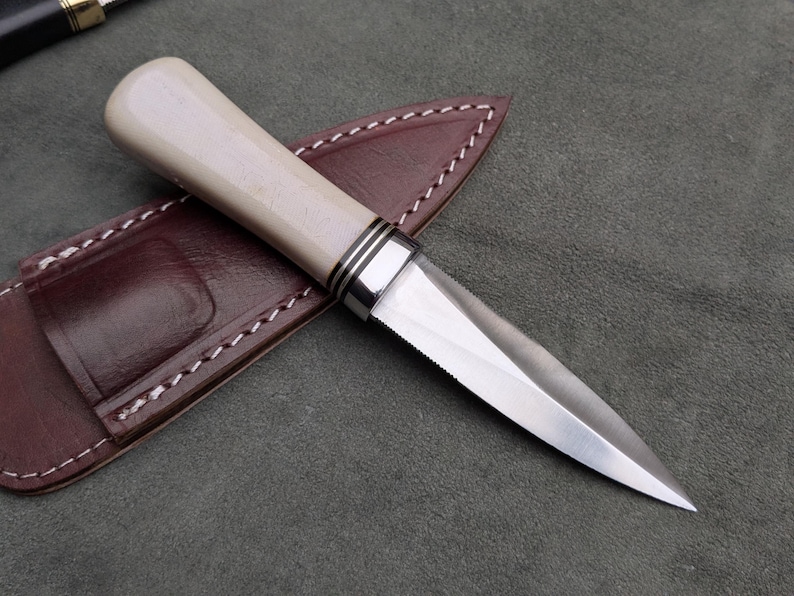 Pu&ograve; includere: Un coltello artigianale con un manico colorato chiaro e una lama argentata, appoggiato su un fodero in pelle marrone. Il manico ha dettagli argentati. Il fodero &egrave; cucito con filo bianco. Il coltello &egrave; progettato per l'uso.