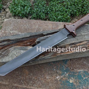 May include: A large, straight-edged knife with a dark, polished wooden handle and a wide, gray blade. The knife is resting on a weathered wooden surface. The text "Haritagestudio" is visible on the blade.