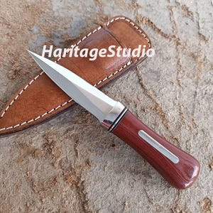 K&ouml;nnte beinhalten: Ein silbernes und braunes Taschenmesser mit einem Holzgriff und einer Lederscheide. Das Messer hat eine scharfe Klinge und eine spitze Spitze.