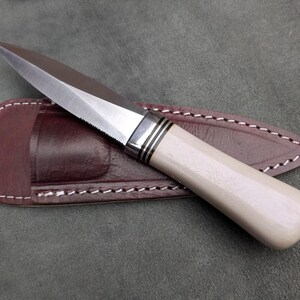Pu&ograve; includere: Un coltello fatto a mano con una lama in acciaio lucido e un manico color crema. Il coltello &egrave; appoggiato su un fodero in pelle marrone con cuciture bianche. La lama ha un bordo seghettato vicino all'impugnatura.