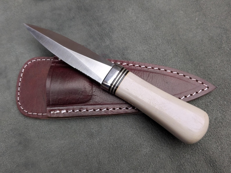 Pu&ograve; includere: Un coltello fatto a mano con una lama in acciaio lucido e un manico color crema. Il coltello &egrave; appoggiato su un fodero in pelle marrone con cuciture bianche. La lama ha un bordo seghettato vicino all'impugnatura.