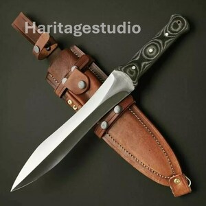 May include: A silver knife with a black and white patterned handle and a brown leather sheath. The knife is sharp and has a pointed tip. The sheath has a belt loop and a snap closure. The text "Haritagestudio" is visible on the sheath.