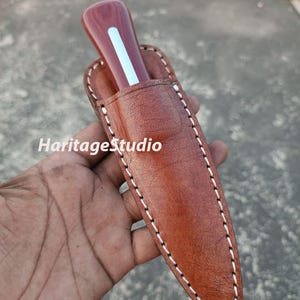 K&ouml;nnte beinhalten: Eine braune Ledertasche f&uuml;r ein Messer mit wei&szlig;en N&auml;hten. Die Tasche hat eine spitze Spitze und eine Klappe, die mit einem Druckknopf befestigt wird. Ein Messer mit Holzgriff befindet sich in der Tasche.