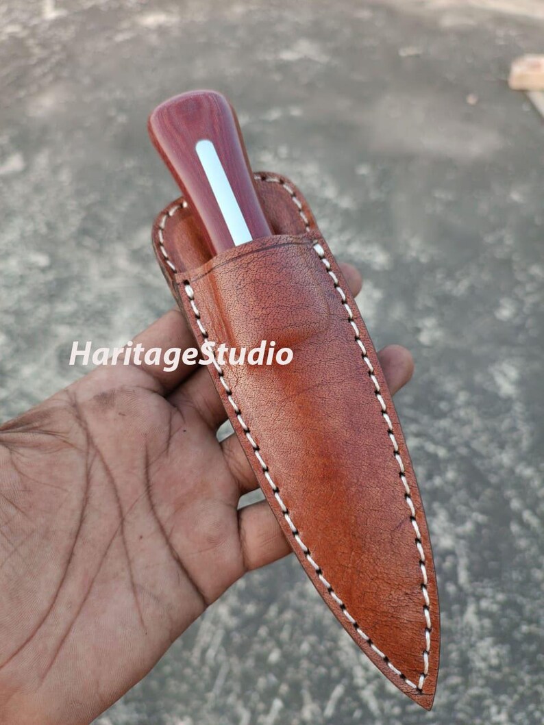 K&ouml;nnte beinhalten: Eine braune Ledertasche f&uuml;r ein Messer mit wei&szlig;en N&auml;hten. Die Tasche hat eine spitze Spitze und eine Klappe, die mit einem Druckknopf befestigt wird. Ein Messer mit Holzgriff befindet sich in der Tasche.