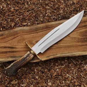 May include: A fixed-blade knife featuring a polished steel blade with a central fuller, a brass crossguard, and a textured brown antler handle. The knife is positioned diagonally across a piece of wood, showcasing its sharp point and edge.