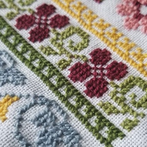 May include: Close-up of a white fabric with a colorful embroidered design. The design features a repeating pattern of flowers in shades of pink, red, green, and blue.