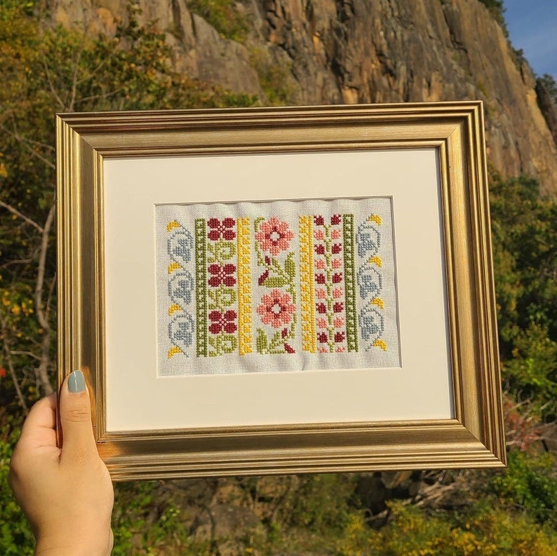 May include: A framed cross-stitch embroidery with a floral design. The embroidery features a repeating pattern of flowers, leaves, and swirls in shades of red, pink, yellow, green, and blue.