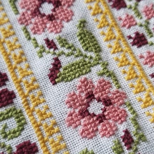 May include: Close-up of a white fabric with a cross-stitch pattern. The pattern features pink, yellow, green, and burgundy flowers and leaves.