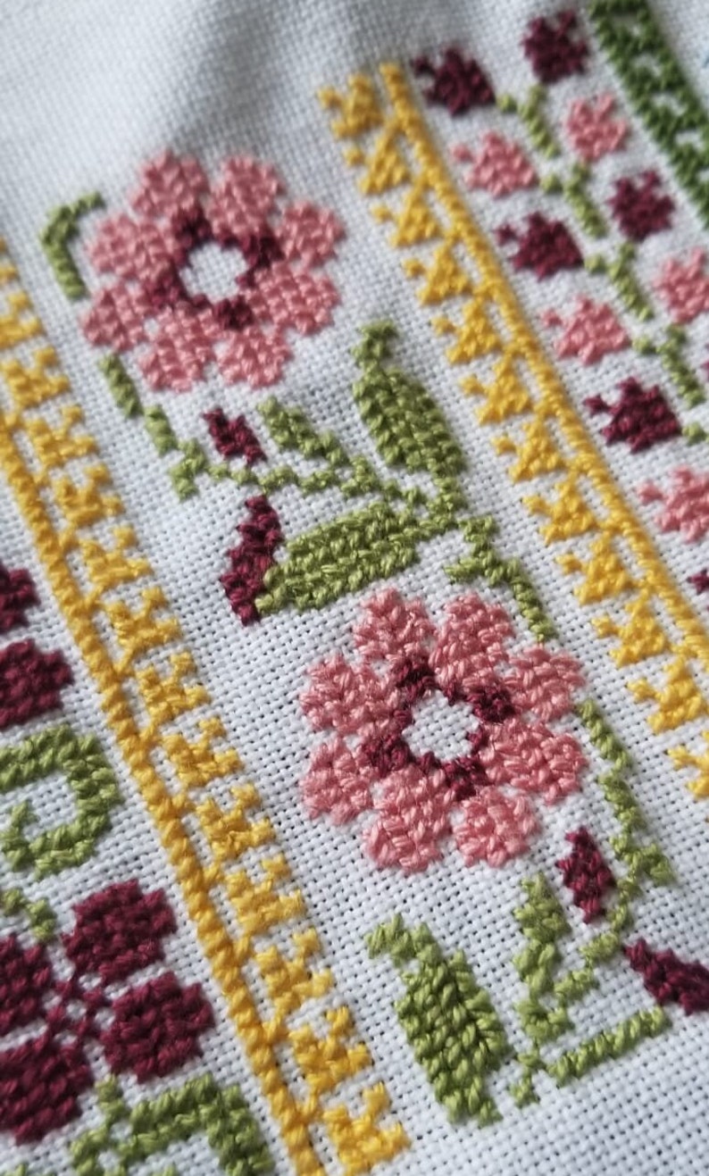 May include: Close-up of a white fabric with a cross-stitch pattern. The pattern features pink, yellow, green, and burgundy flowers and leaves.