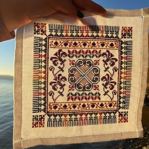 Peut inclure: Un carr&eacute; de lin blanc avec un motif brod&eacute; color&eacute; pr&eacute;sentant un motif central de quatre volutes entrelac&eacute;es. La bordure est d&eacute;cor&eacute;e d'un motif r&eacute;p&eacute;titif de feuilles stylis&eacute;es et de formes g&eacute;om&eacute;triques dans des tons de rouge, vert, marron et or.