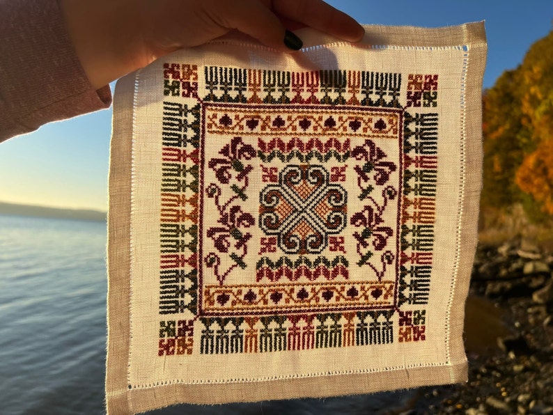 Peut inclure: Un carr&eacute; de lin blanc avec un motif brod&eacute; color&eacute; pr&eacute;sentant un motif central de quatre volutes entrelac&eacute;es. La bordure est d&eacute;cor&eacute;e d'un motif r&eacute;p&eacute;titif de feuilles stylis&eacute;es et de formes g&eacute;om&eacute;triques dans des tons de rouge, vert, marron et or.