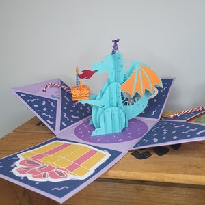 May include: A pop-up birthday card with a turquoise dragon wearing a party hat and holding a cake with a lit candle. The card is open, showing a gift box illustration and a purple and blue background.