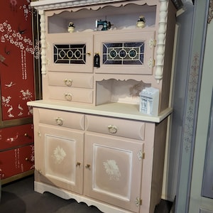 Könnte beinhalten: Ein hellrosa Schrank mit weißer Ober- und Unterseite. Der Schrank hat unten zwei Türen und zwei Schubladen. Der obere Teil hat zwei Türen mit Buntglasfenstern. Dekorative florale Akzente und Beschläge vervollständigen den Look.