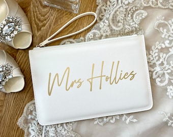 May include: White clutch bag with a gold zipper and a gold inscription that reads "Mrs. Hollies".