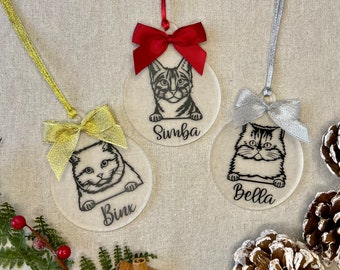 May include: Three circular frosted acrylic ornaments featuring black line art illustrations of cats. Each ornament displays a cat name: Binx, Simba, or Bella. The ornaments are topped with gold, red, and silver ribbons and matching bows.