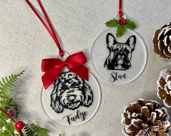 May include: Two clear, round Christmas ornaments with black dog portraits and names. One has a red bow and the name "Fudge", the other has a holly accent and the name "Stan". Both have red ribbons for hanging. Pine cones and greenery are in the background.