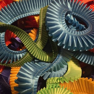 May include: A close-up of colorful, pleated satin ribbons in shades of blue, green, orange, and yellow. The ribbons are arranged in circular and wavy patterns, creating a textured and vibrant display. The ribbons are likely used for crafting or decorative purposes.