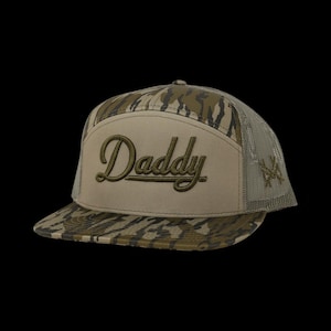 May include: A baseball cap featuring a tan front panel with the word Daddy embroidered in dark green script. The brim and side panels display a camouflage pattern in shades of brown and green. The back panels consist of a tan mesh material.