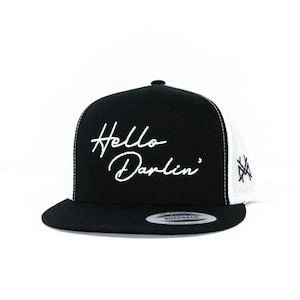 May include: A black and white snapback cap featuring the white embroidered text Hello Darlin' on the front panel. The hat has a black brim, a black front panel, and white mesh side and back panels with a small black embroidered logo on the side.