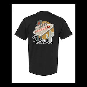 May include: A black short-sleeve t-shirt featuring a graphic on the back. The design includes a retro-style sign with the text I'M THE PROBLEM, a yellow sun, a saguaro cactus, and two white dice. The graphic uses red, white, and yellow colors.