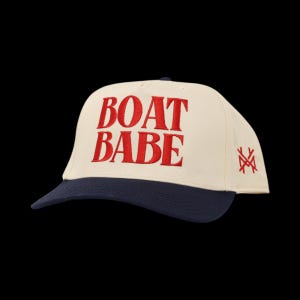 May include: Cream-colored baseball cap with a navy blue brim. The words "BOAT BABE" are embroidered in red on the front. A red monogram is embroidered on the side. The hat is set against a black background.