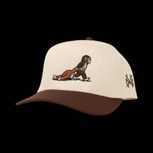 May include: A beige and brown baseball cap featuring an embroidered cartoon monkey carrying another monkey. The cap has a brown brim and top button, with a logo on the side.