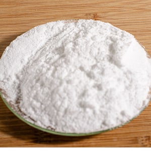 May include: A close-up shot of a white, powdery substance in a small, round, clear dish with a green rim. The dish sits on a wooden surface with a natural grain pattern. The powder appears finely ground and textured.