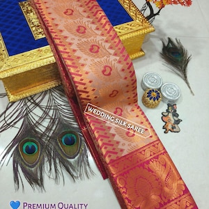 May include: A pink and orange silk saree with a gold border and peacock design. The saree is folded and displayed on a white surface with a wooden box and peacock feathers.