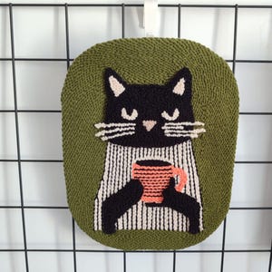 Può includere: Un arazzo da parete fatto a mano a forma di gatto verde oliva. Il gatto nero tiene in mano una tazza color corallo. Il gatto ha i baffi bianchi e indossa un maglione a righe. L'arazzo da parete è realizzato in filato strutturato.