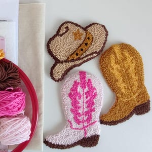 May include: Three colourful, textured, handmade punch needle designs: a cowboy hat, a cowboy boot, and a cowgirl boot. The hat is tan with a brown brim and star details. The boots are pink and yellow with brown accents. A red embroidery hoop and yarn are in the background.