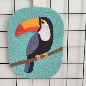 May include: A turquoise, oval-shaped wall hanging featuring a toucan bird design. The toucan has a large orange and red beak, white face, black body, and gray wings, perched on a brown branch. The background is a textured turquoise.