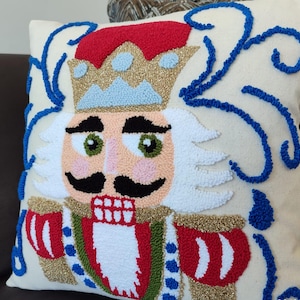 May include: A white pillow with a colorful embroidered nutcracker design. The nutcracker is wearing a red hat, a gold crown, and a red and white striped jacket. The background is white with blue swirls.