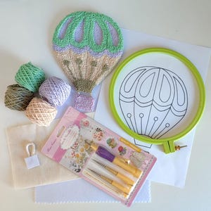 Op de afbeelding: Een heteluchtballon-ponsnaaldset met groene, paarse en bruine borduurgaren, een houten borduurring, een naaldgereedschap en een stuk stof.