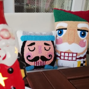 Può includere: Due cuscini di peluche con volti di schiaccianoci ricamati. Un cuscino ha un cappello blu e un baffo, l'altro ha un cappello verde e una barba bianca. I cuscini sono su una superficie di legno.